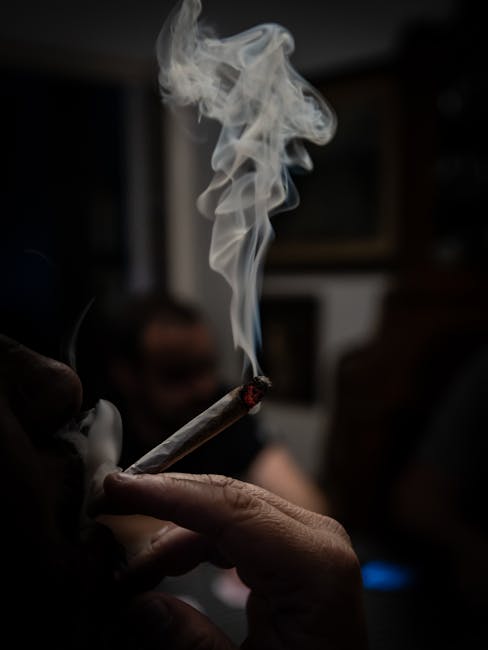 A close-up shot of a hand holding a cigarette with smoke trails in a dim setting.